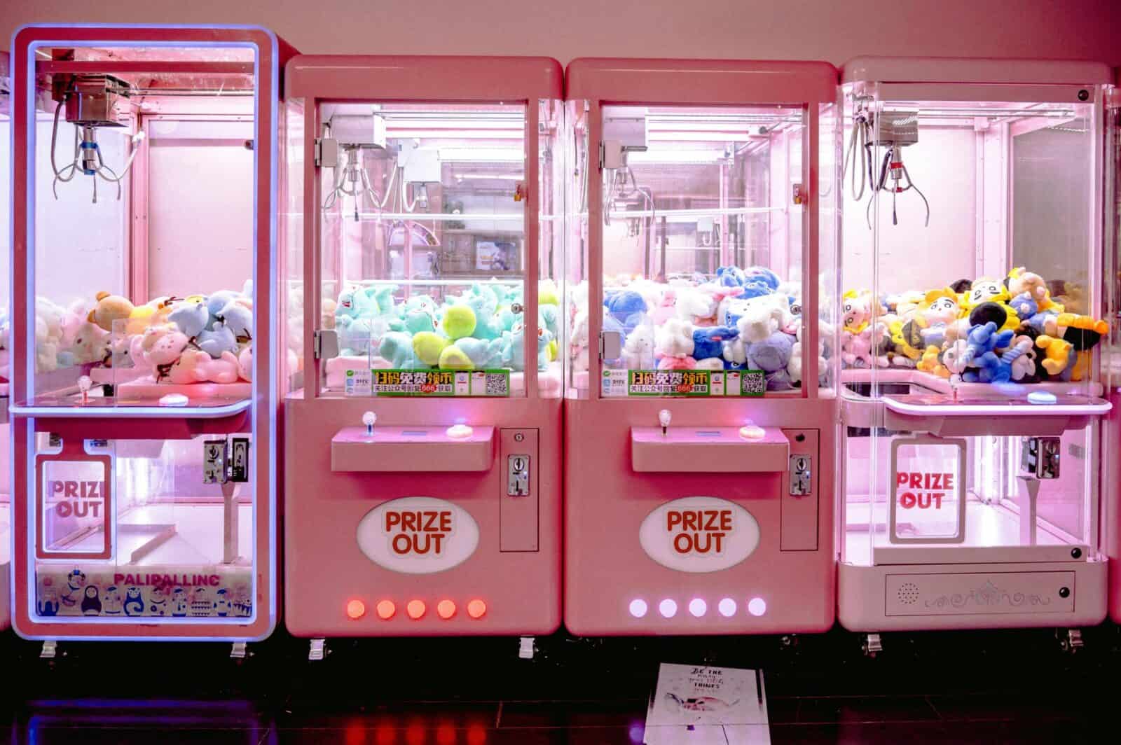 Rows of illuminated pink claw machines filled with vibrant soft toys indoors.